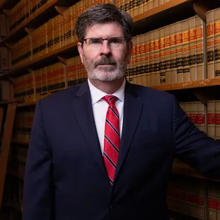 Middle-aged man in dark suit and red striped tie standing in front of law bookshelves.