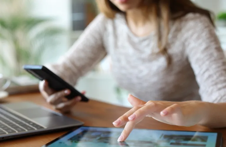 Woman using a smartphone and touching a tablet screen at a wooden desk.