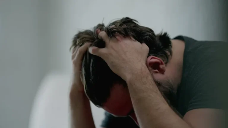 Man with dark hair and beard holding his head in both hands, appearing distressed.