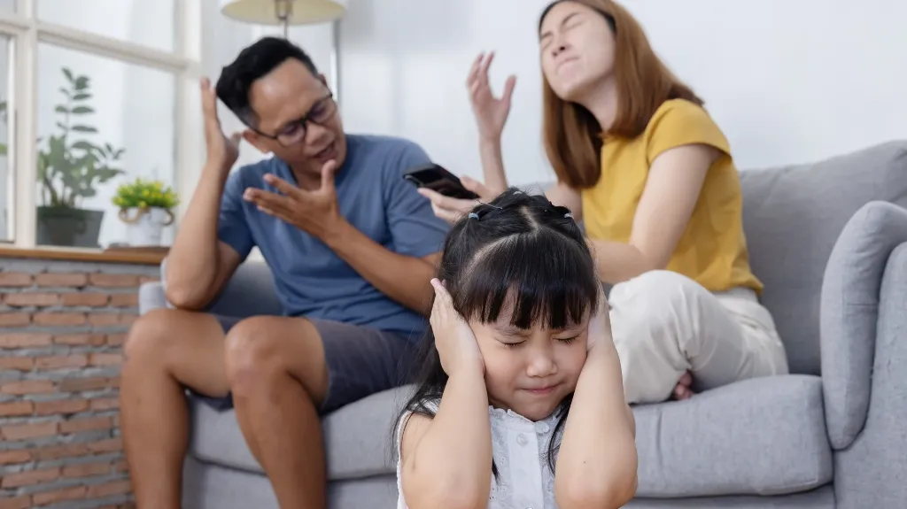 Young girl covering ears in front of arguing man and woman seated on gray sofa.